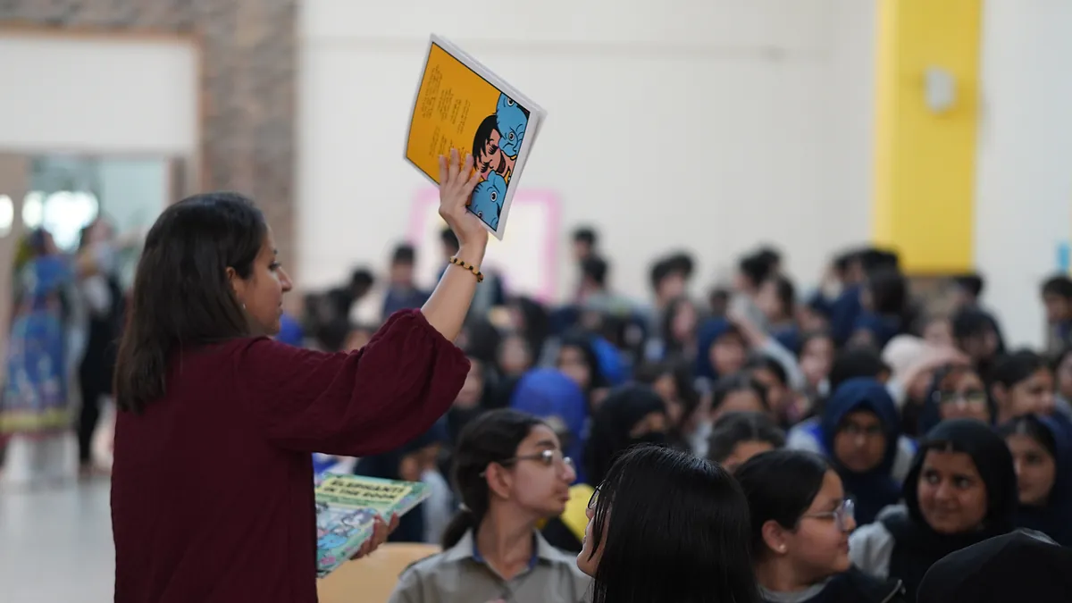 A pupil holds a Killing Cancer Kindly comic high above a hall full of seated schoolchildren.