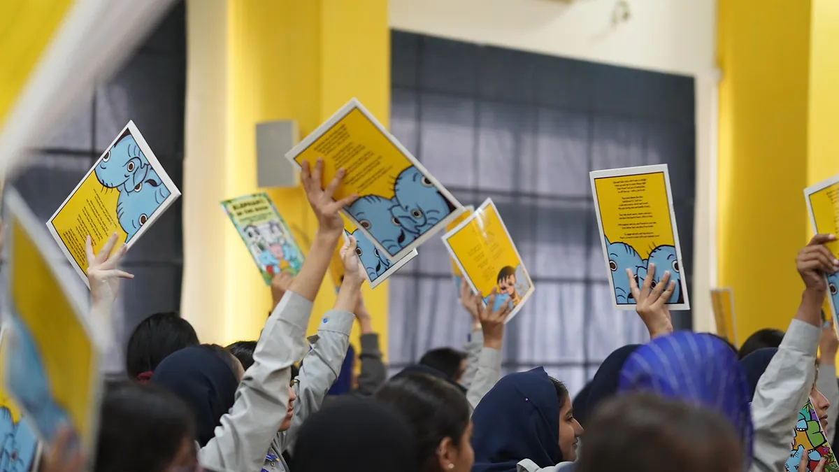 Hands reaching up from a packed school hall, each holding a Killing Cancer Kindly prevention comic.